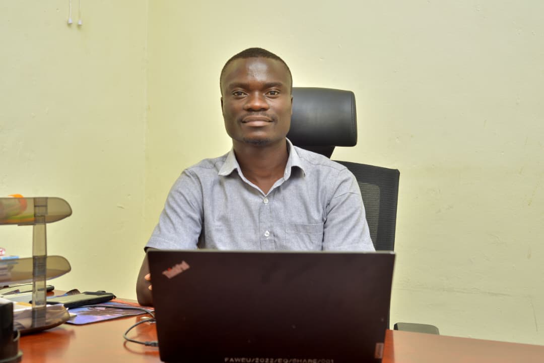 Man seated at a desk with an open laptop, facing the camera in a simple office setting.